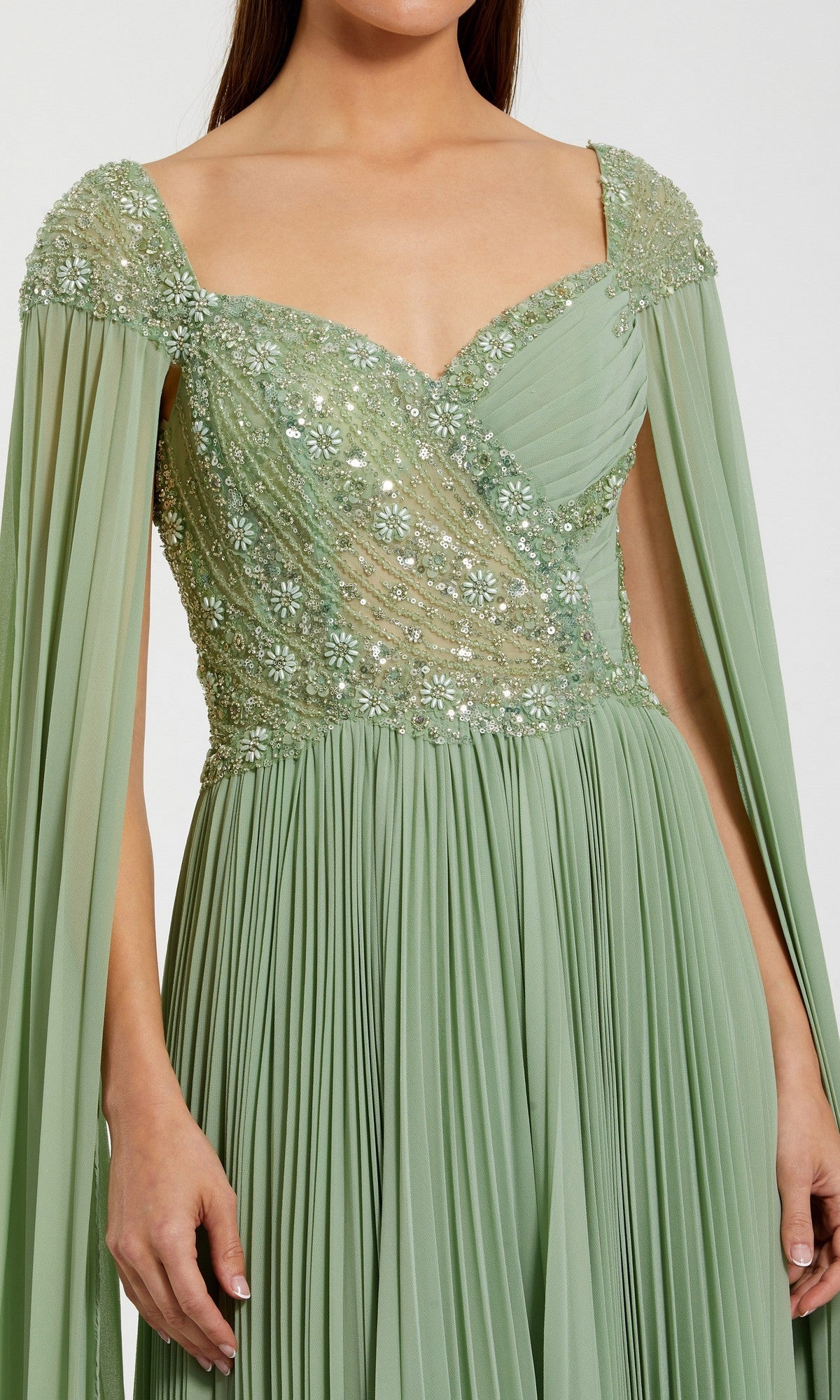 A woman wears the Mac Duggal 37034 long formal dress in light green, featuring a pleated chiffon skirt, floral embellished bodice, and draped cape-like sleeves.